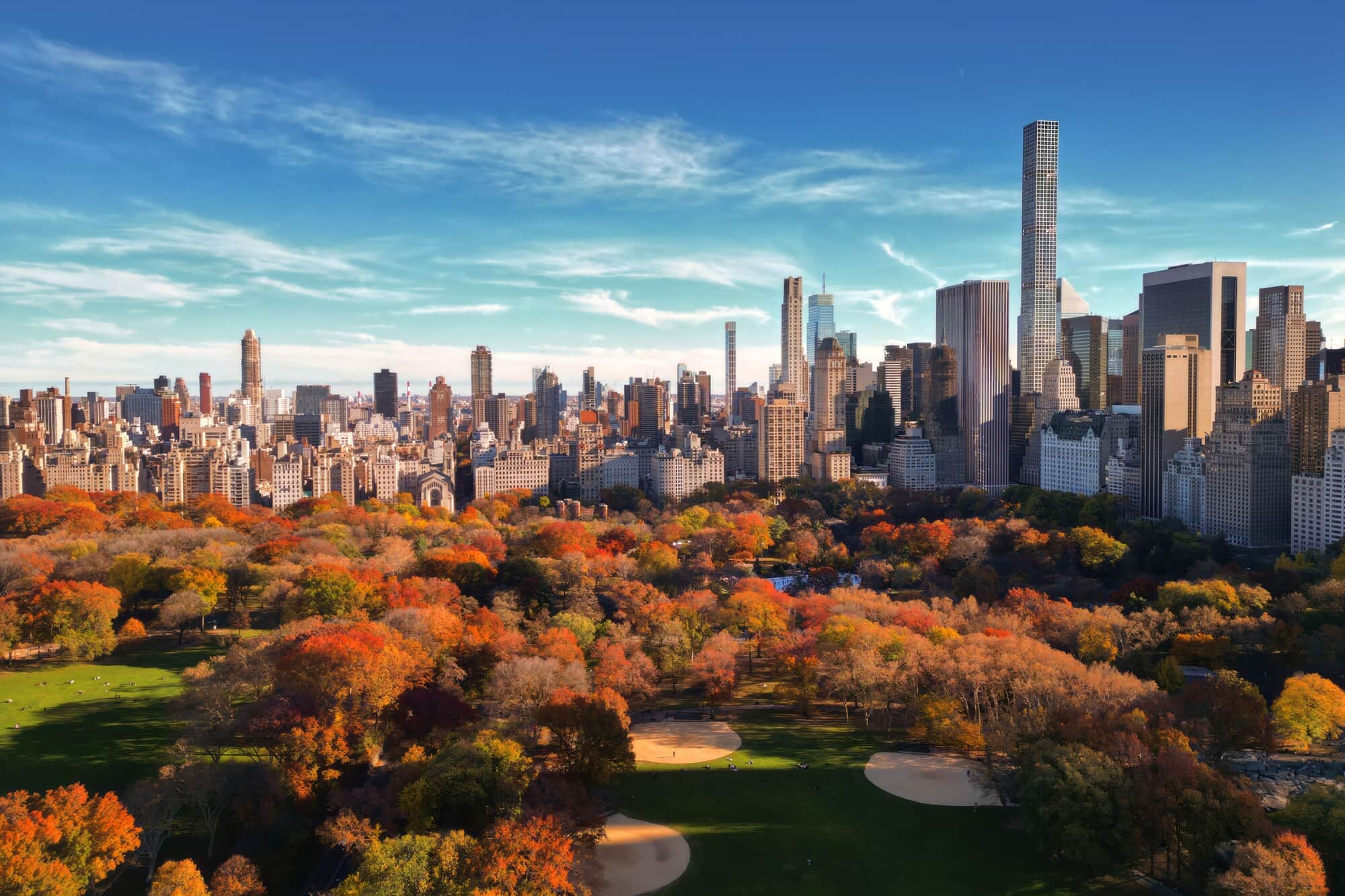 Central Park autumn scene: colorful trees, calm water, and iconic New York City skyscrapers under a cloudy sky.